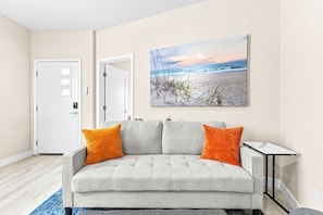 A living room with a grey sofa, two orange cushions, a small table, and a beach scene painting on the wall. - New 2BR Home Minutes to Downtown SD (San Diego)