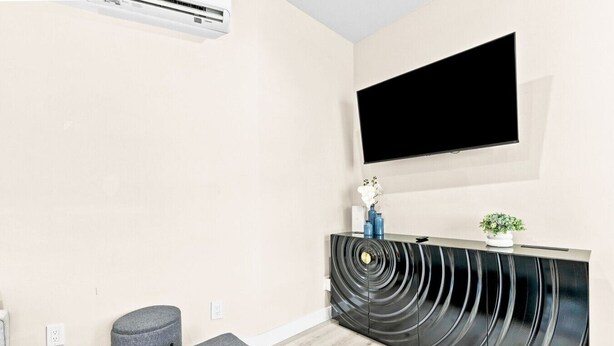 A modern living room with a flat-screen TV mounted on the wall, a black curved entertainment unit, a grey ottoman, and a small potted plant. - New 2BR Home Minutes to Downtown SD (San Diego)