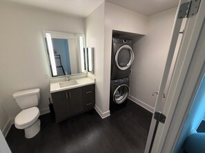 Ein kleines Badezimmer mit Toilette, dunklem Boden, Waschbecken mit Spiegel und gestapelter Waschmaschine und Trockner.
