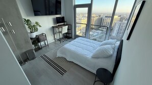 Ein modernes Schlafzimmer mit einem großen Bett, einem Schreibtisch, einem Stuhl, einem Fernseher und Blick auf die Stadt durch große Fenster.