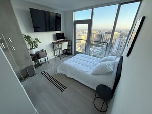 Ein modernes Schlafzimmer mit einem großen Bett, einem Schreibtisch, einem Stuhl, einem Fernseher und Blick auf die Stadt durch große Fenster.