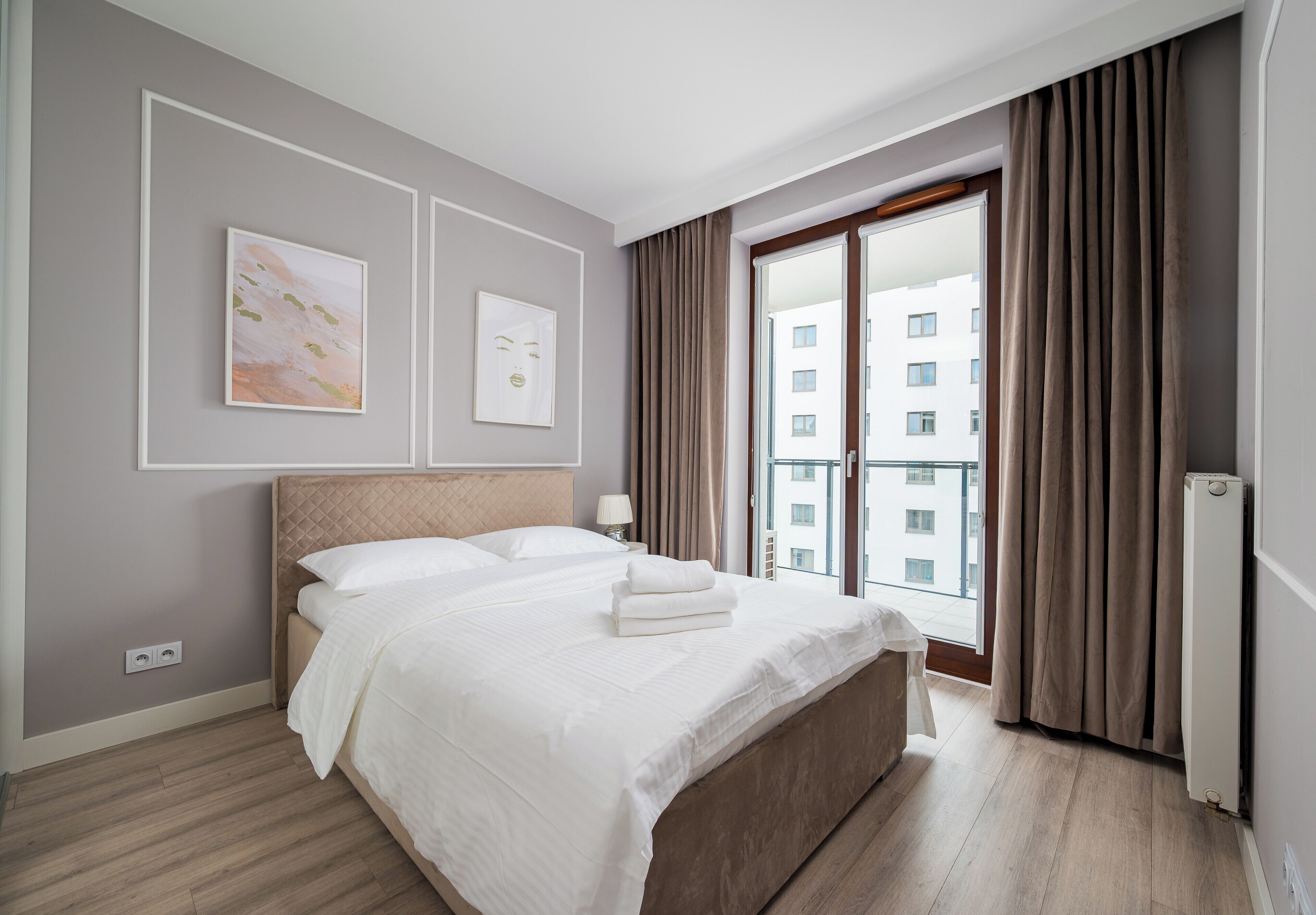 Ein modernes Hotelzimmer mit einem großen Bett, Blick auf die Stadt durch eine Schiebetür und einem kleinen Nachttisch mit Lampe.