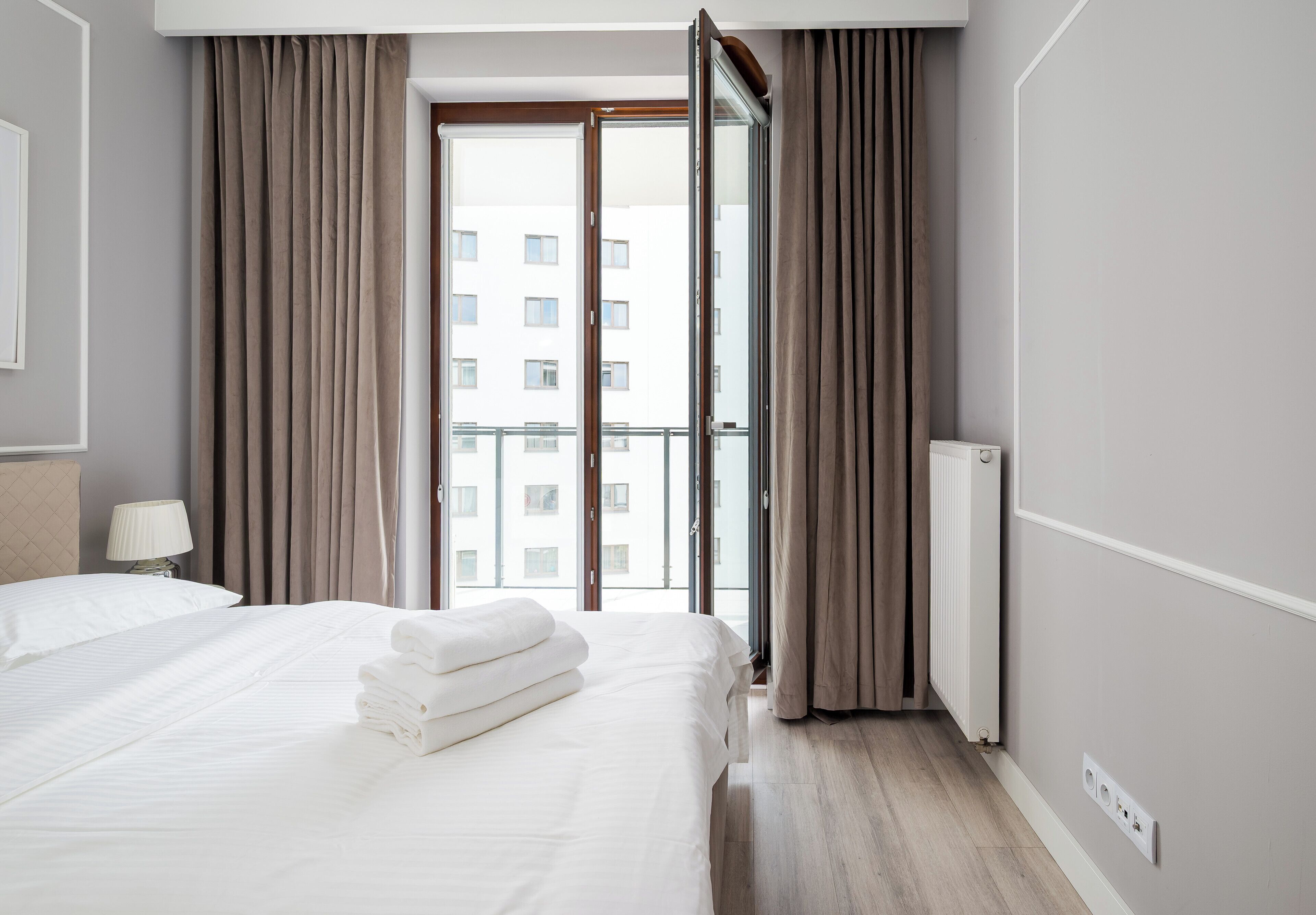 A hotel room with a bed, a lamp, a radiator, and a view of a building through a glass door.