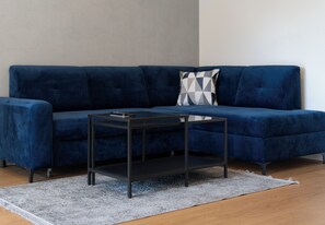 En blå soffa med en geometrisk mönstrad kudde, ett svart soffbord och en grå matta på ett trägolv.