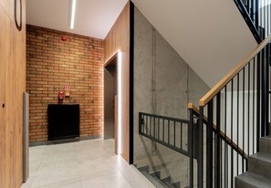 A modern interior with a brick wall, a black radiator, a wooden reception desk, and a staircase with a glass railing. - Portowa Riverside Apartment (Kraków)