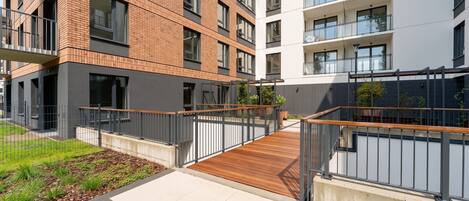 A modern apartment building with a wooden deck, metal railings, and a landscaped garden area.