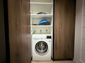 A compact laundry room with a front-loading washing machine, a shelf above it, and a blue appliance on the top shelf. - Madalińskiego Prime Studio 3B (Wrocław)