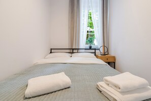A neatly made bed with pillows and two folded towels, a bedside table with a lamp, and a window with curtains. - Krzywa Harmony Apartment (Katowice)