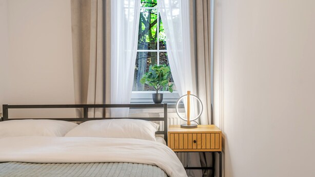A bedroom with a bed, a bedside table with a mirror, and a window with curtains. - Krzywa Harmony Apartment (Katowice)