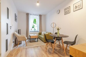 A modern living room with a sofa, a dining table with chairs, a small side table, and a window with curtains. - Krzywa Harmony Apartment (Katowice)