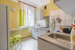 A compact kitchen with a gas stove, a sink, and a window with curtains. There are two stools and a clock on the wall. - Powstańców Central Apartment (Katowice)