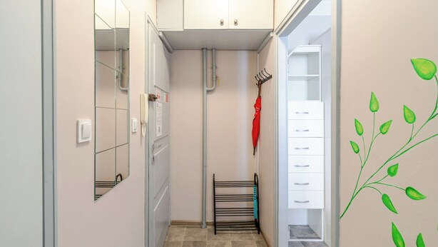 En smal hall med ett klinkergolv, en klädhängare med ett rött paraply, ett vitt skåp med l ådor och en vägg med en illustration av en grön växt.