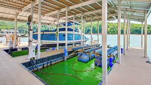 Un muelle techado con un bote azul, agua verde y un paisaje boscoso de fondo.