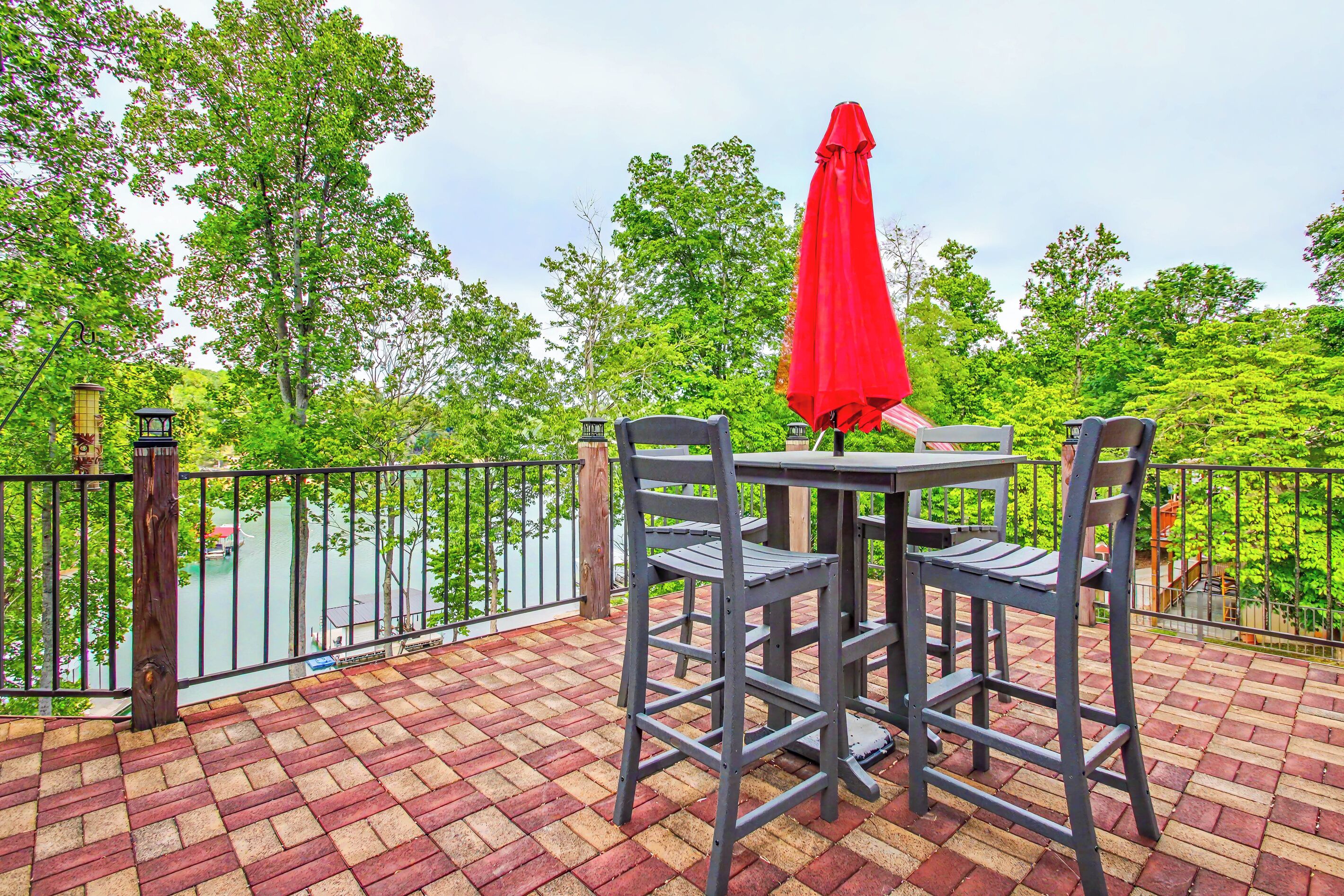 Une terrasse avec une table et des chaises, un parasol rouge, et une vue sur une étendue d’eau et des arbres.