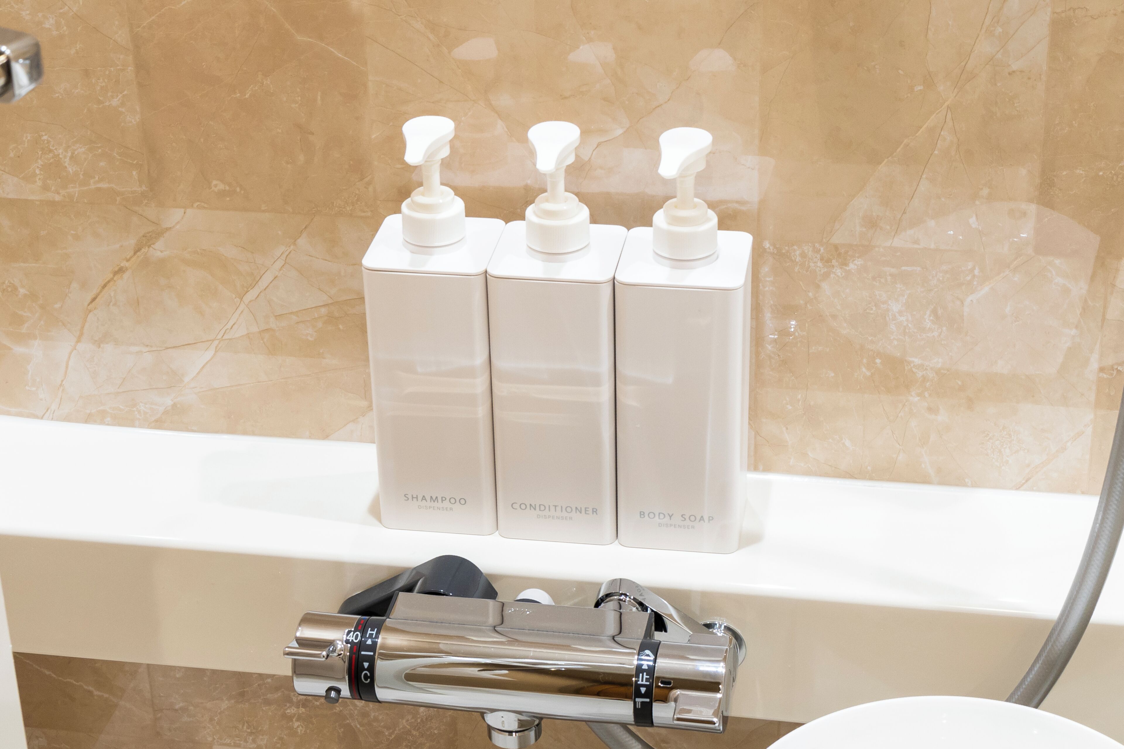 A bathroom with a marble wall, a white shelf holding three white dispensers labeled 'SHAMPOO DISPENSER,' 'CONDITIONER DISPENSER,' and 'BODY SOAP DISPENSER.'