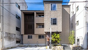 A modern apartment building with a mix of brick and stone facades, multiple balconies, and a small garden area with steps leading to the entrance.