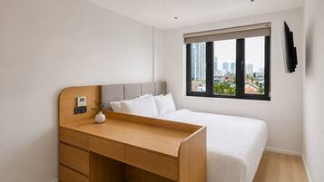A bedroom with a bed, a wooden desk, a small white vase, a window with a city view, and a TV mounted on the wall.