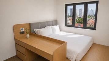Une chambre à coucher avec un lit, un bureau en bois, un vase avec une plante et une fenêtre donnant sur la ville.
