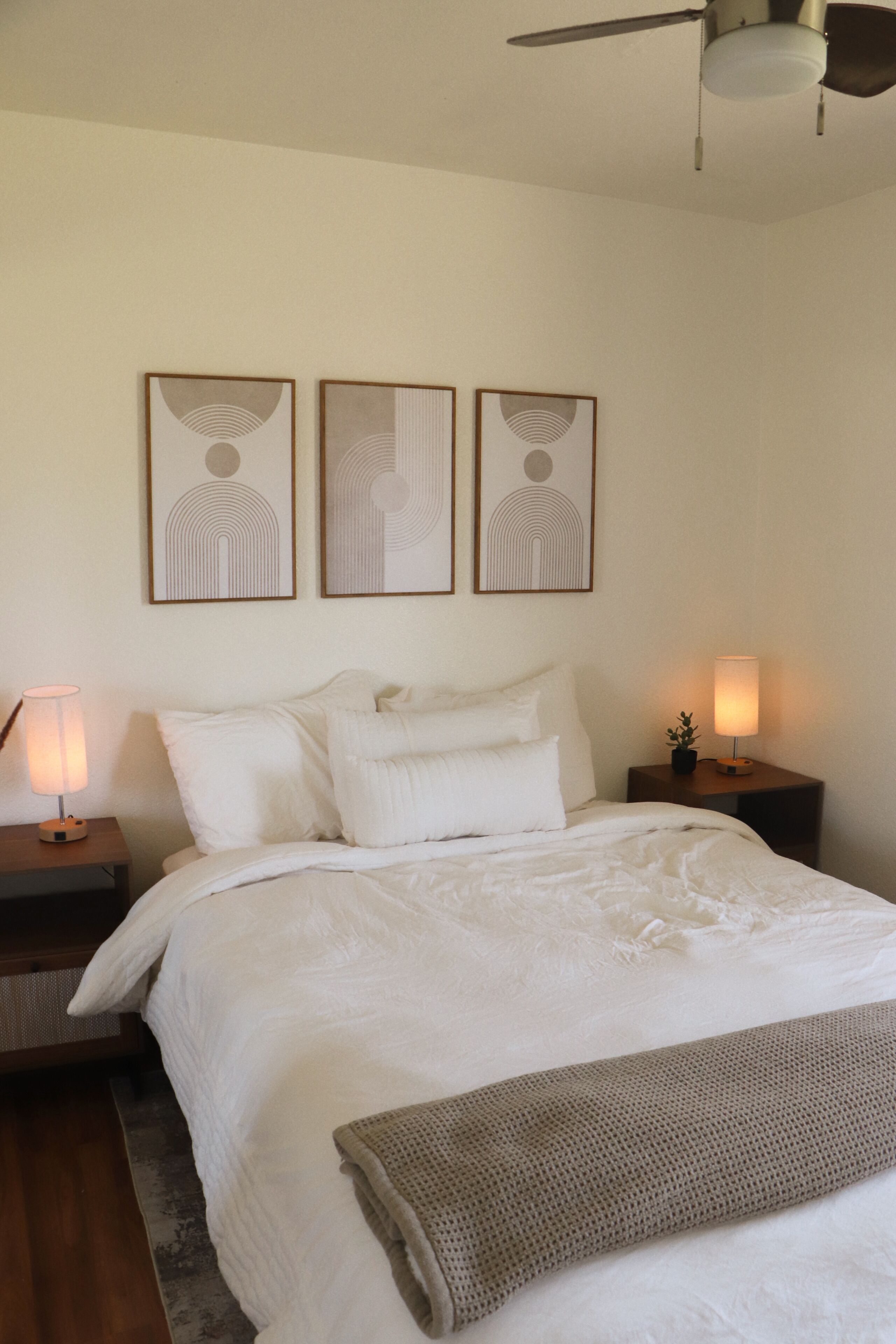 Une chambre à coucher comprenant un lit, deux tables de chevet, un ventilateur de plafond et trois œuvres d’art accrochées au mur.