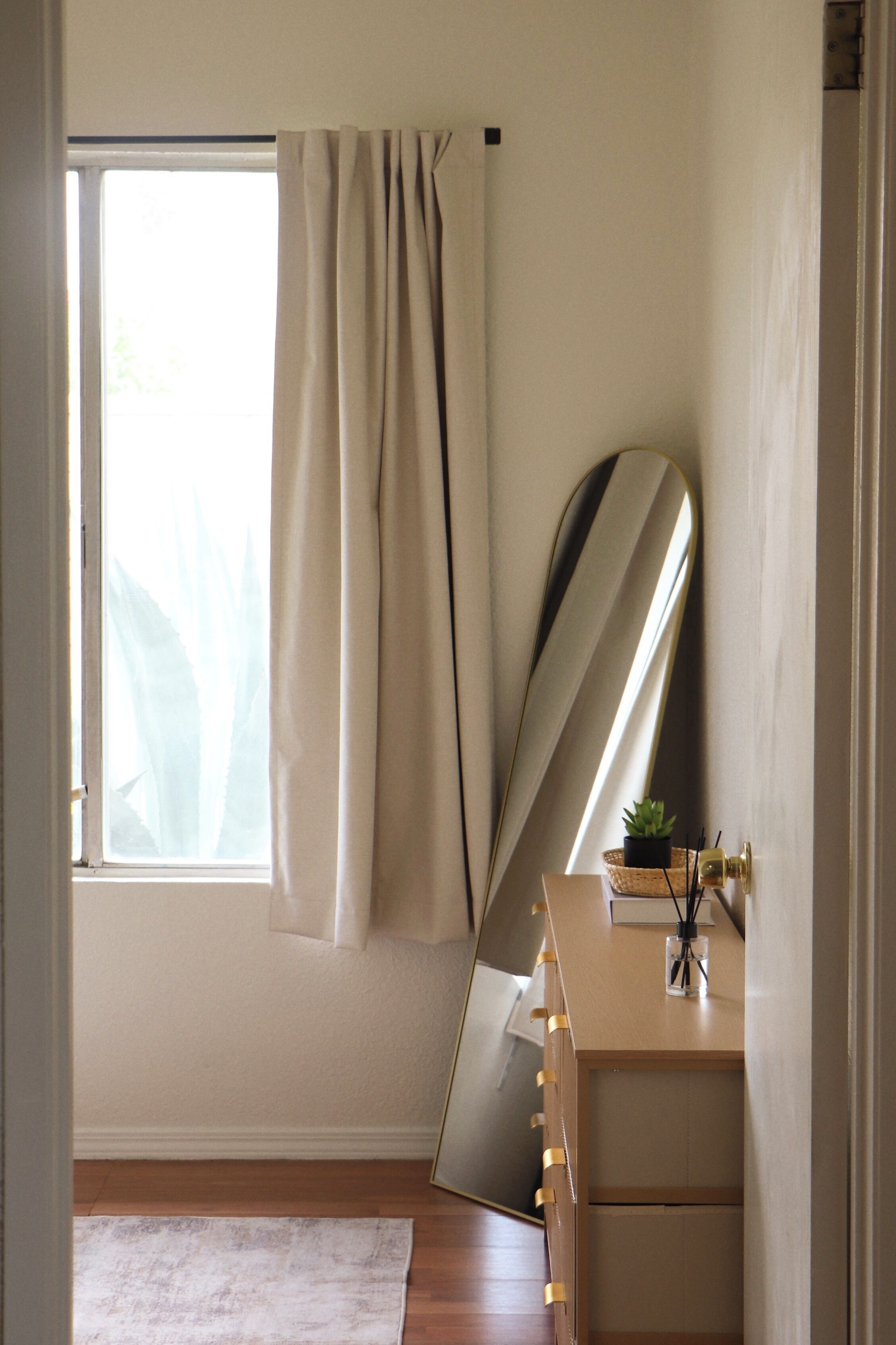 Une pièce avec une grande fenêtre, un miroir, une commode en bois et une petite plante.