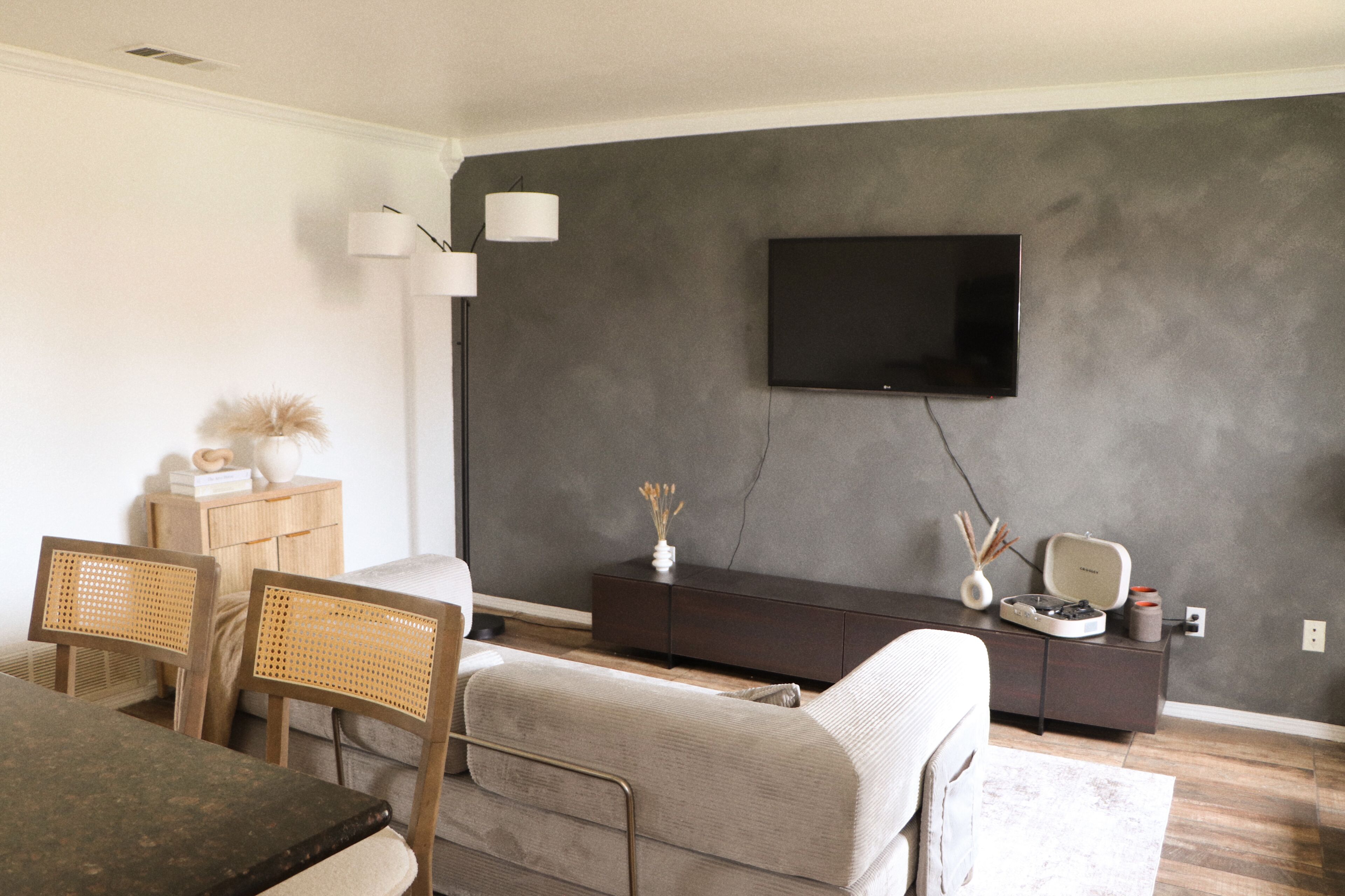 A living room with a flat-screen TV mounted on the wall, a dark-colored entertainment unit, a wooden side table with decorative items, and a sofa with a throw blanket.