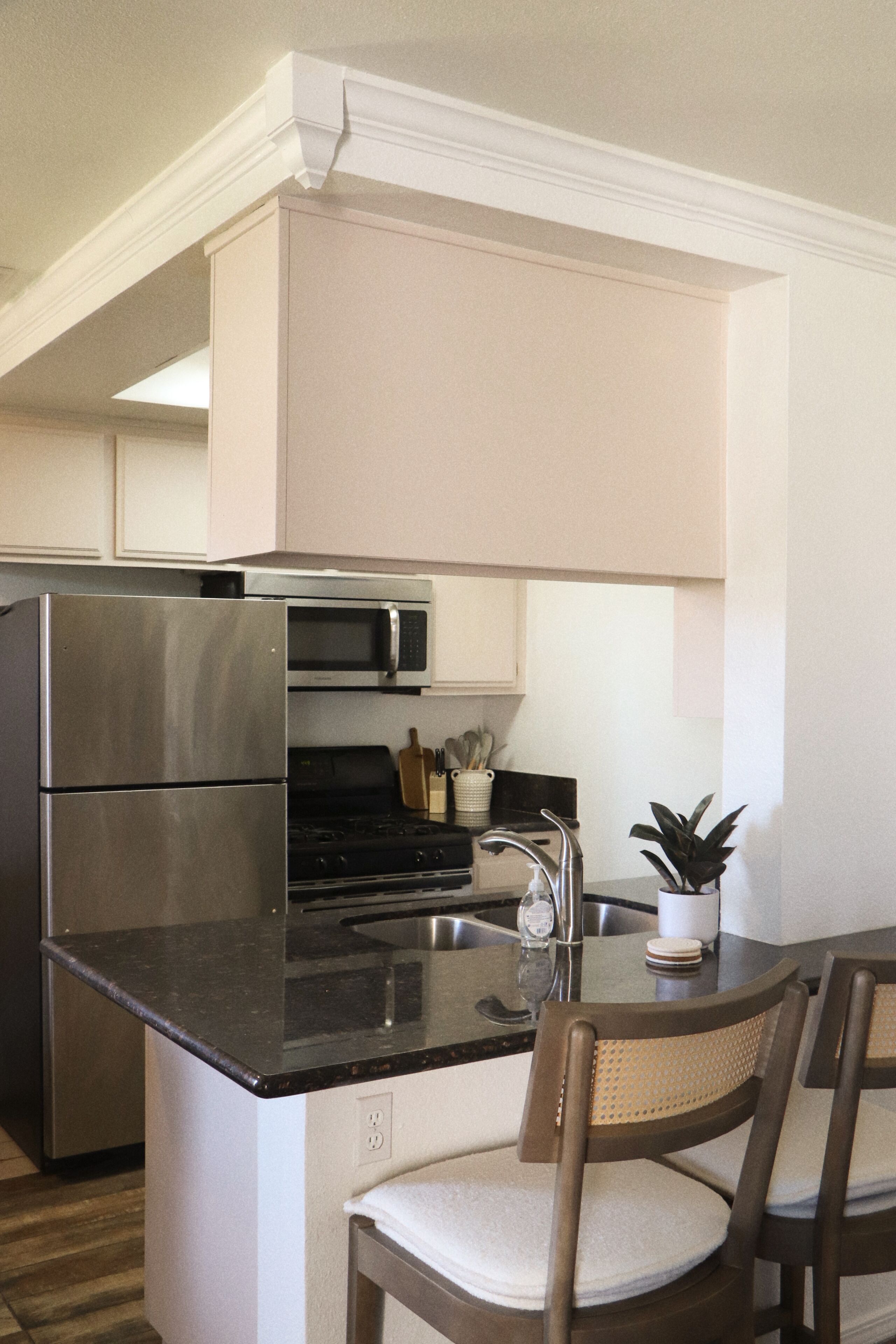 A compact kitchen with a refrigerator, microwave, stove, and a sink. There is a chair with a cushion and a potted plant on the counter.