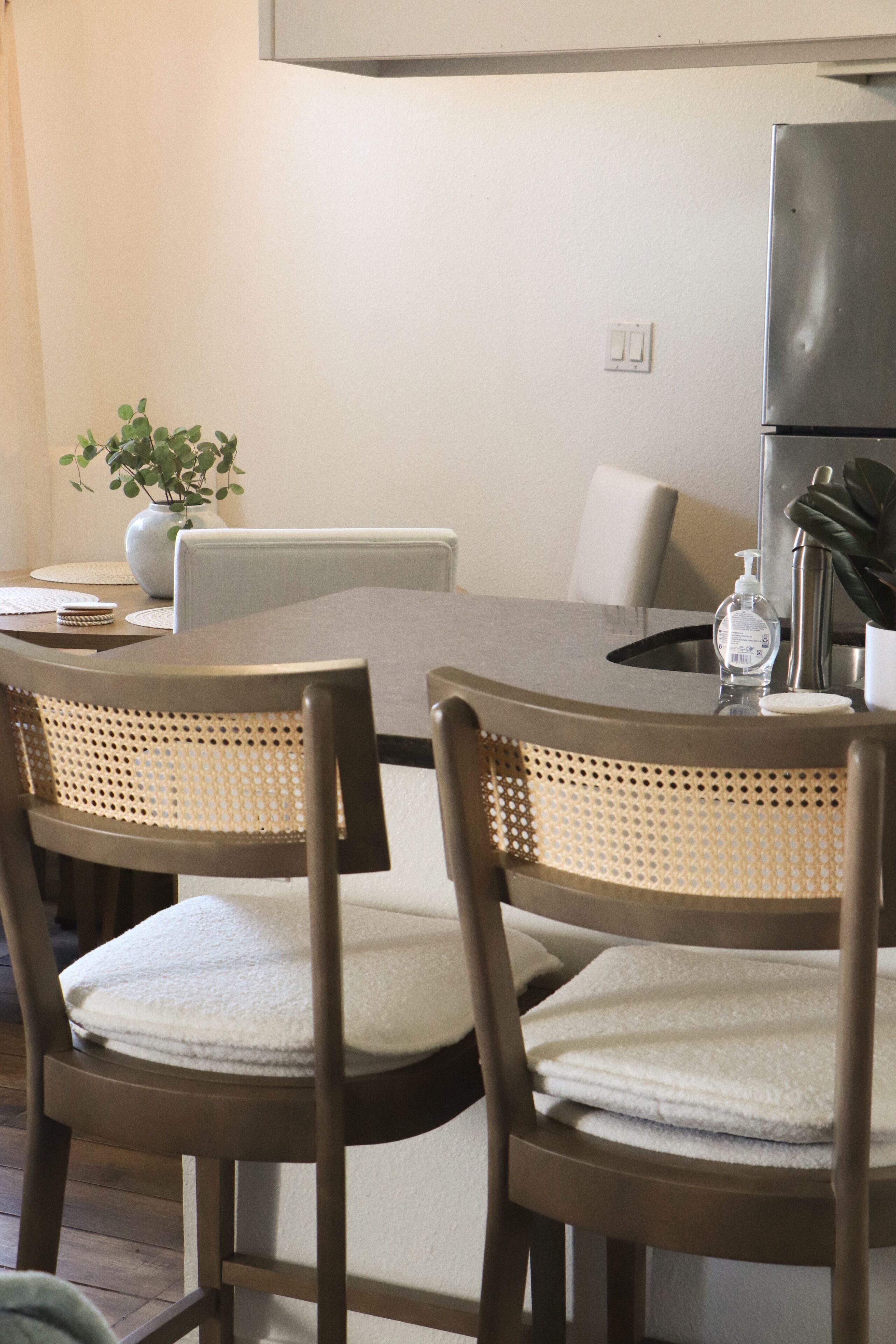 Un coin repas comprenant une table ronde, deux chaises à dossier en osier, un vase avec une plante et une bouteille d’eau.