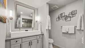 A bathroom with a large mirror, a sink, a toilet, and a shower. There are towels hanging on the wall and a painting above the mirror. - Trail's End - 22-zone3197 3 Bedroom Condo (Estes Park)