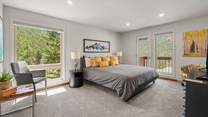 A modern bedroom with a large bed, two bedside lamps, a chair, and a view of the outdoors through large windows. - Trail's End - 22-zone3197 3 Bedroom Condo (Estes Park)