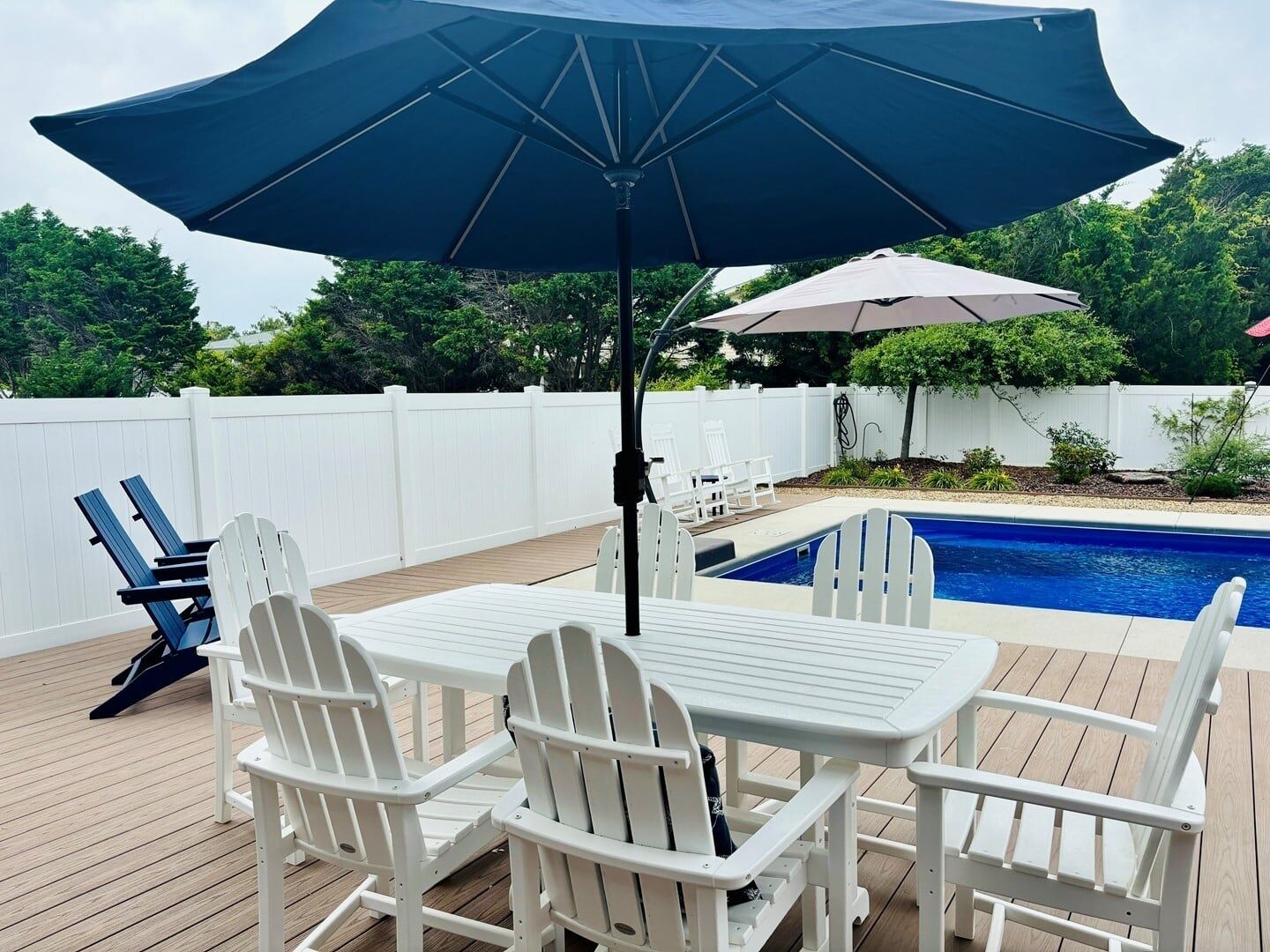 An outdoor deck with a pool, white Adirondack chairs, and a table with an umbrella.