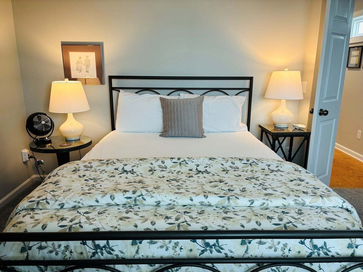 A bed with a floral bedspread, two bedside lamps, a framed picture on the wall, and a door to the right.
