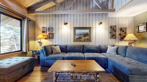 A cozy living room with a sectional sofa, a wooden coffee table, and a large window with a view of trees. - Seasons 4 #104 2 Bedroom Condo (Mammoth Lakes)