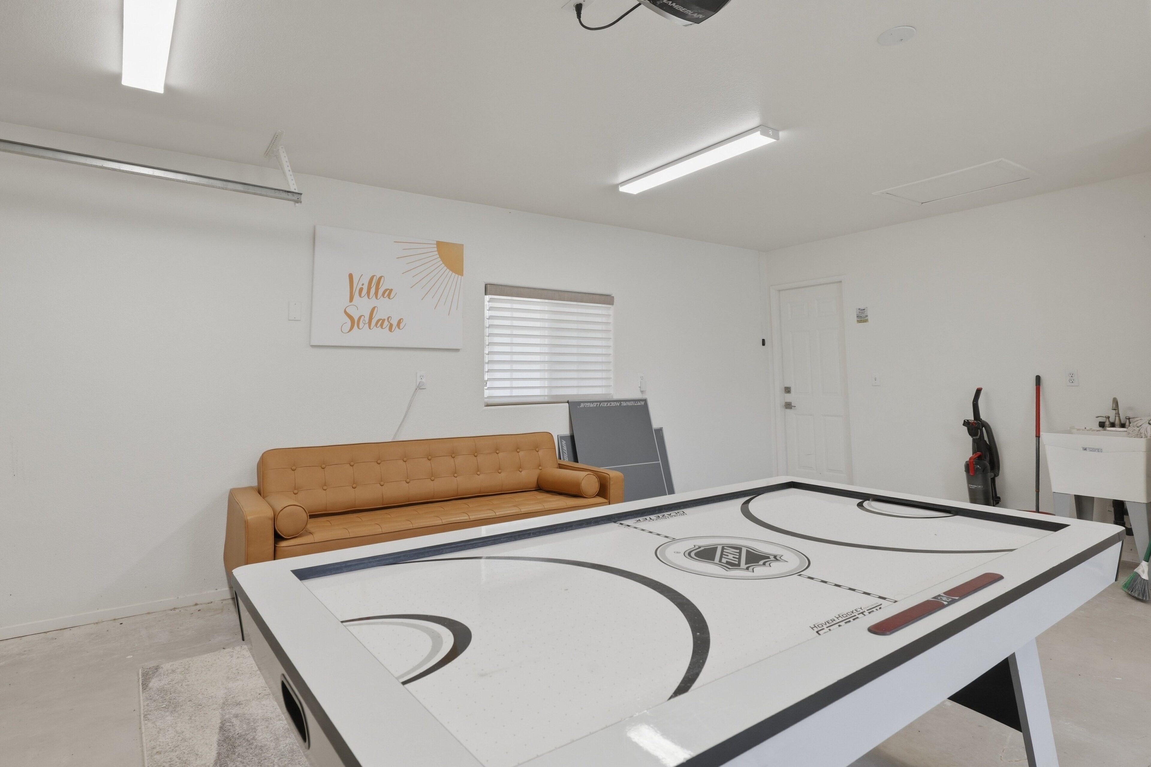 A room with a white table hockey table, a brown sofa, a window with blinds, and a wall with a 'Villa Soleil' sign.