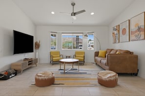 A living room with a flat-screen TV, two yellow chairs, a brown sofa, a ceiling fan, and a large window with a view of the outside. - Joshua Tree El Oasis 3br Retreat For 6 Guests 3 Bedroom Home (Joshua Tree)