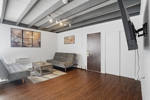 Ein modernes Wohnzimmer mit grauem Sofa, einem kleinen Couchtisch und einem grauen Sessel. An der Wand hängen zwei gerahmte Kunstwerke, die Decke hat sichtbare Balken, und ein Flachbildfernseher ist an der Wand angebracht.