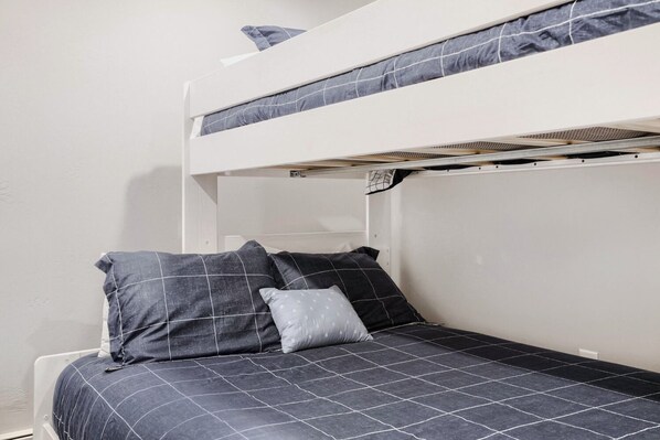 Uma cama beliche com colchão e travesseiros, uma parede branca e uma porta.