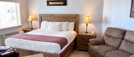 A hotel room with a bed, two bedside tables with lamps, a brown leather sofa, a window with blinds, and a ceiling fan.