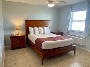 Une chambre à coucher avec un lit en bois, une table de chevet, une lampe, une fenêtre avec des stores et un ventilateur de plafond.
