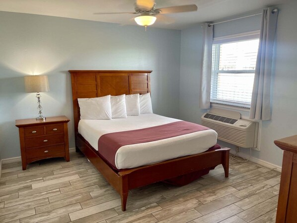 A bedroom with a wooden bed, a nightstand, a lamp, a ceiling fan, a window with blinds, and an air conditioning unit. - Beachgate Condo Suites And Hotel 517a+517b 2 Bedroom Condo (Port Aransas)