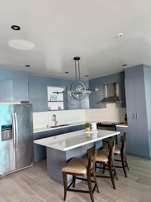 A modern kitchen with a central island, bar stools, and a stainless steel refrigerator. - Ross Rosarito (Playas de Rosarito)