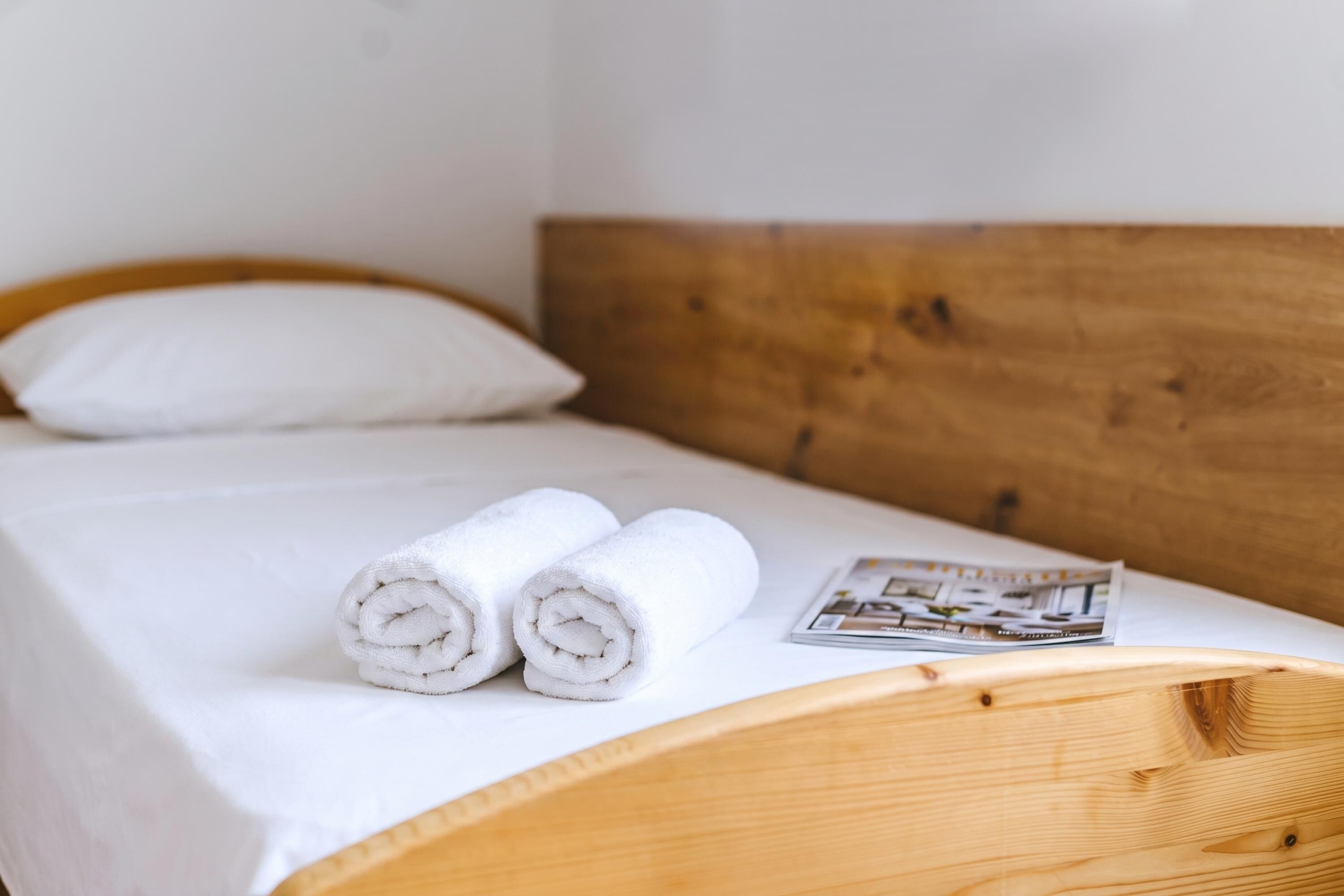 Un lit avec une tête de lit en bois, une literie blanche, deux serviettes blanches roulées et un magazine posé sur le lit.