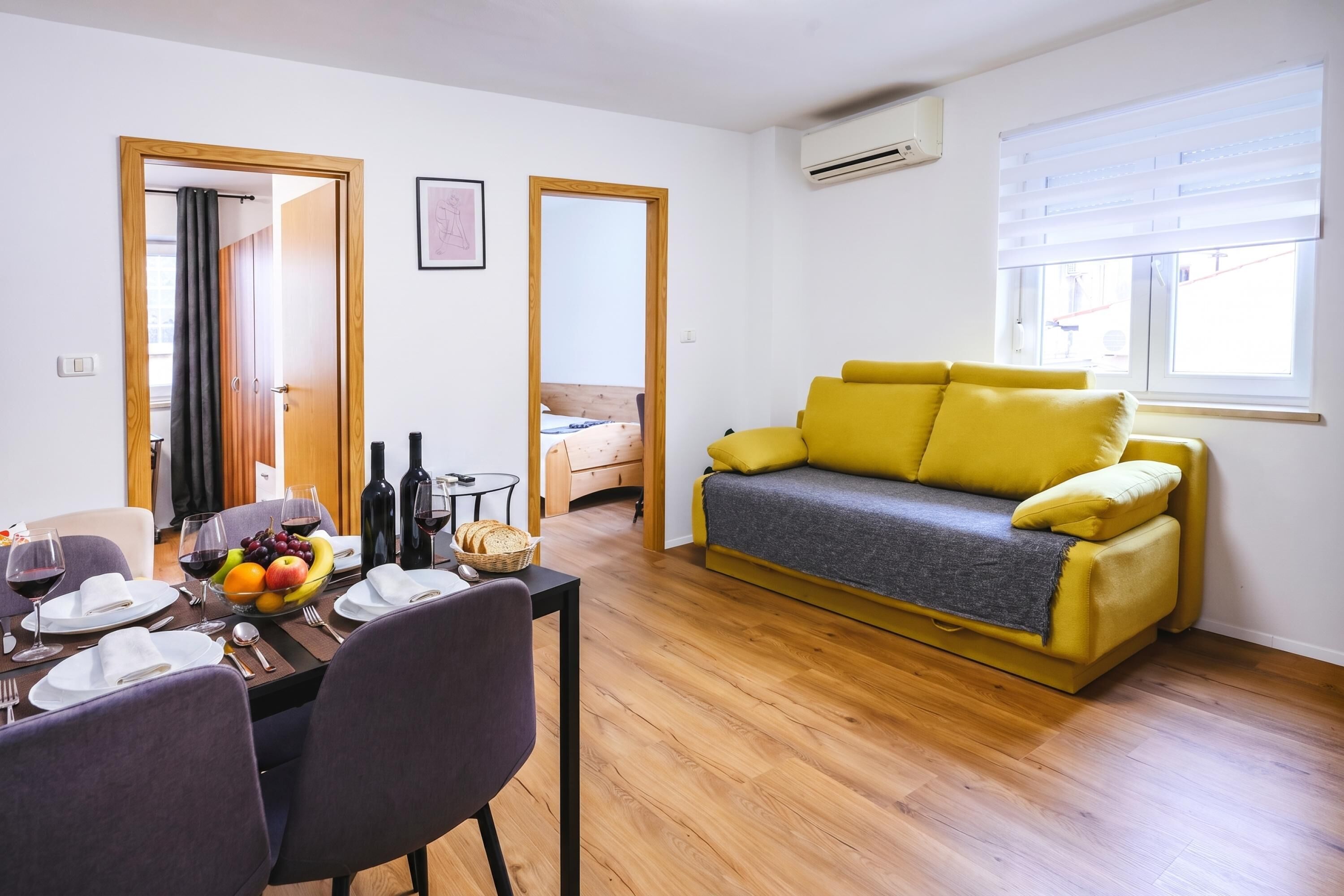A modern living room with a yellow sofa, a dining table set with plates, glasses, and a fruit bowl.
