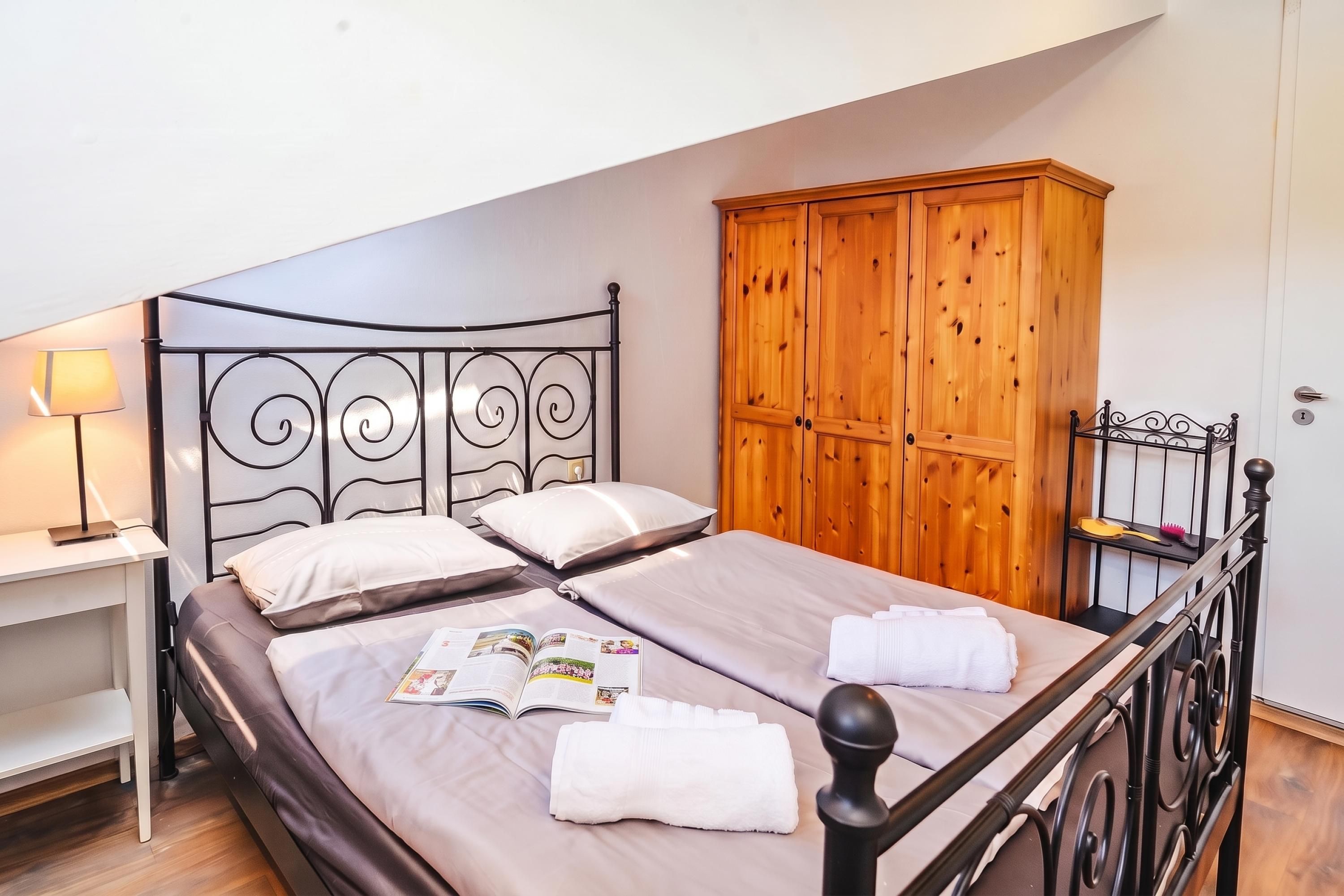 A single bed with a metal frame, a wooden wardrobe, and a bedside table with a lamp.