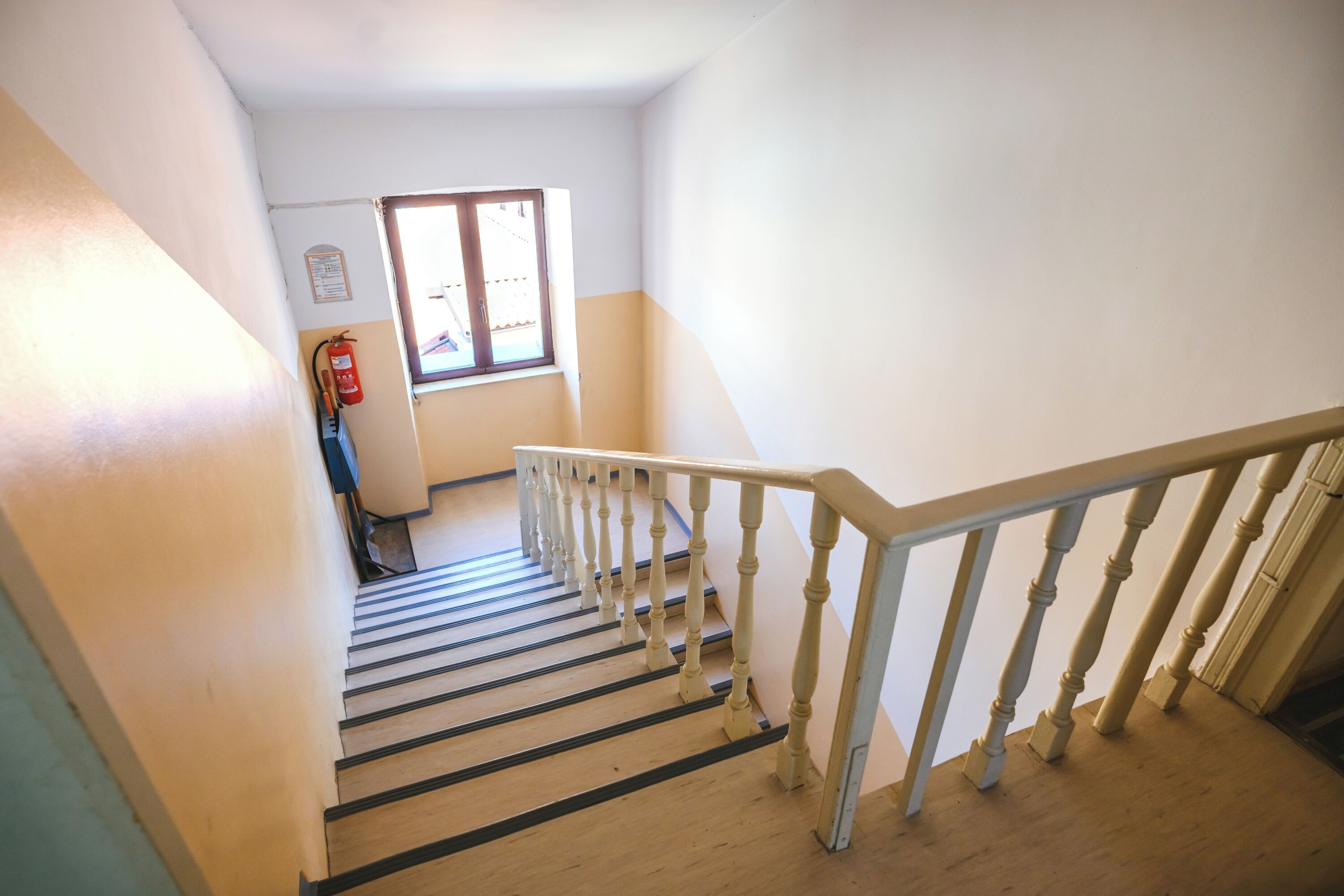 Eine Treppe mit Holzbodenstufen und weißen Geländern, die zu einer Zwischenebene mit einem Fenster und einem an der Wand befestigten Feuerlöscher führt.