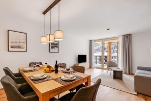 Uma área de jantar moderna com uma mesa de madeira posta para refeição, uma televisão e vista para uma paisagem nevada através de grandes janelas.