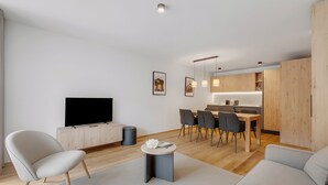En moderne stue med et fladskærms-tv, en sofa, et spiseområde med stole, og et køkken i baggrunden.
