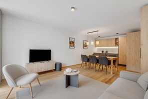 Een moderne woonkamer met een flatscreen televisie, een bank, een eethoek met stoelen en een keuken op de achtergrond.