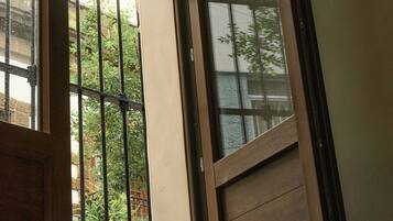 Una habitación con una puerta corredera que da a un balcón, una ventana con rejas y vistas al exterior con vegetación.