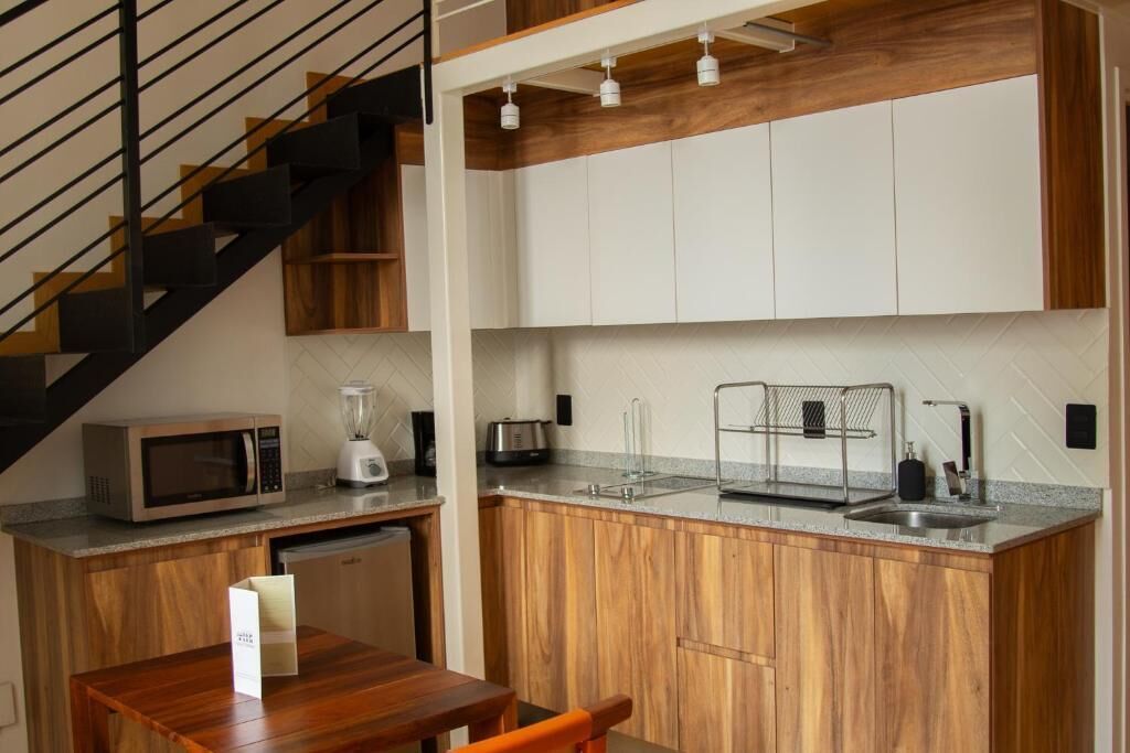 Cocina compacta con microondas, tostadora y licuadora sobre la encimera, armarios de madera, un lavamanos pequeño y una escalera con barandillas negras.