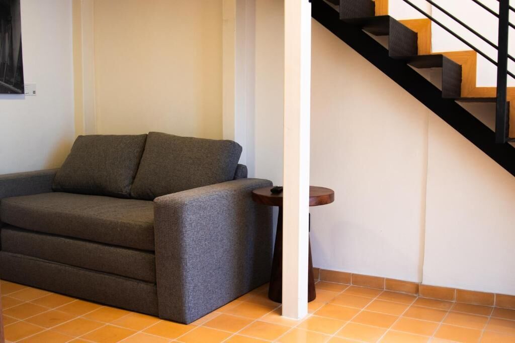 Un espacio de estar con un sofá y una pequeña mesa, una escalera de madera y piso de baldosas.